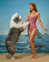 Woman in a pink swimsuit standing on a beach with a dog and dolphins in the background