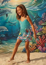 Young girl in a floral dress standing on sand with an underwater mural featuring dolphins and coral in the background.