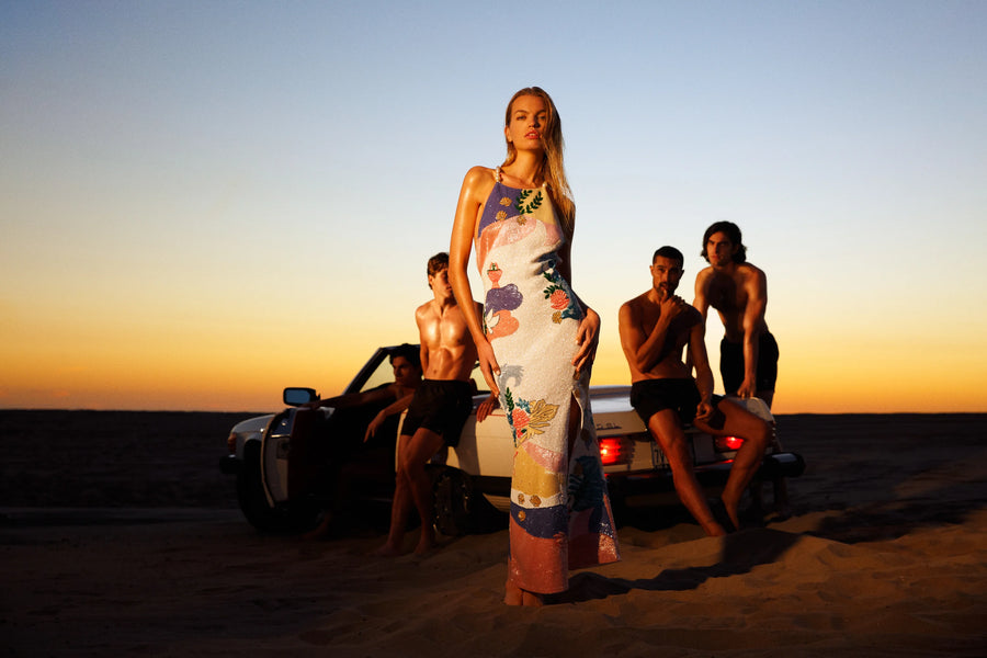 A model wearing a sea shell-inspired halterneck maxi dress with soft pastel hues and intricate beaded detailing. The dress features a figure-skimming silhouette, delicate floral and sea-inspired patterns, and a thigh-high slit for added elegance and movement. The shimmering embellishments add a touch of luxury and coastal charm