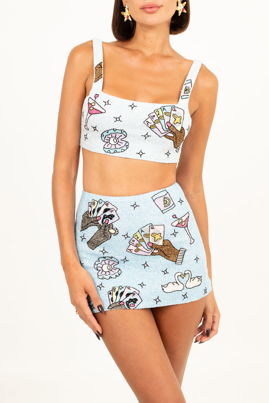 Model wearing a sparkling blue two-piece set with hand-embroidered motifs of poker hands, cocktails, and swans, featuring a crop top with a square neckline and a matching mini skirt.