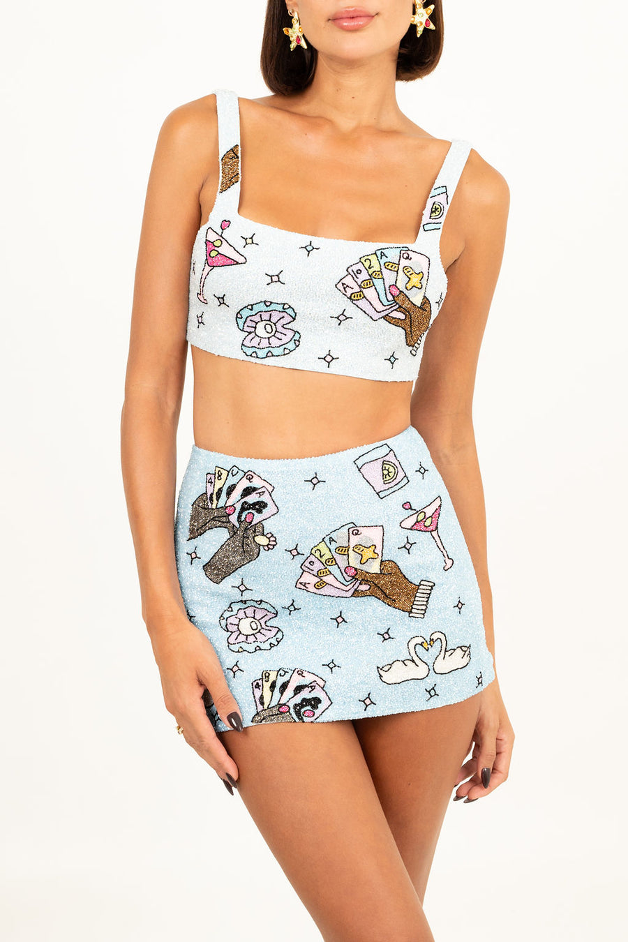 Model wearing a sparkling blue two-piece set with hand-embroidered motifs of poker hands, cocktails, and swans, featuring a crop top with a square neckline and a matching mini skirt.