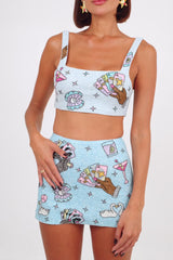 Model wearing a sparkling blue two-piece set with hand-embroidered motifs of poker hands, cocktails, and swans, featuring a crop top with a square neckline and a matching mini skirt.